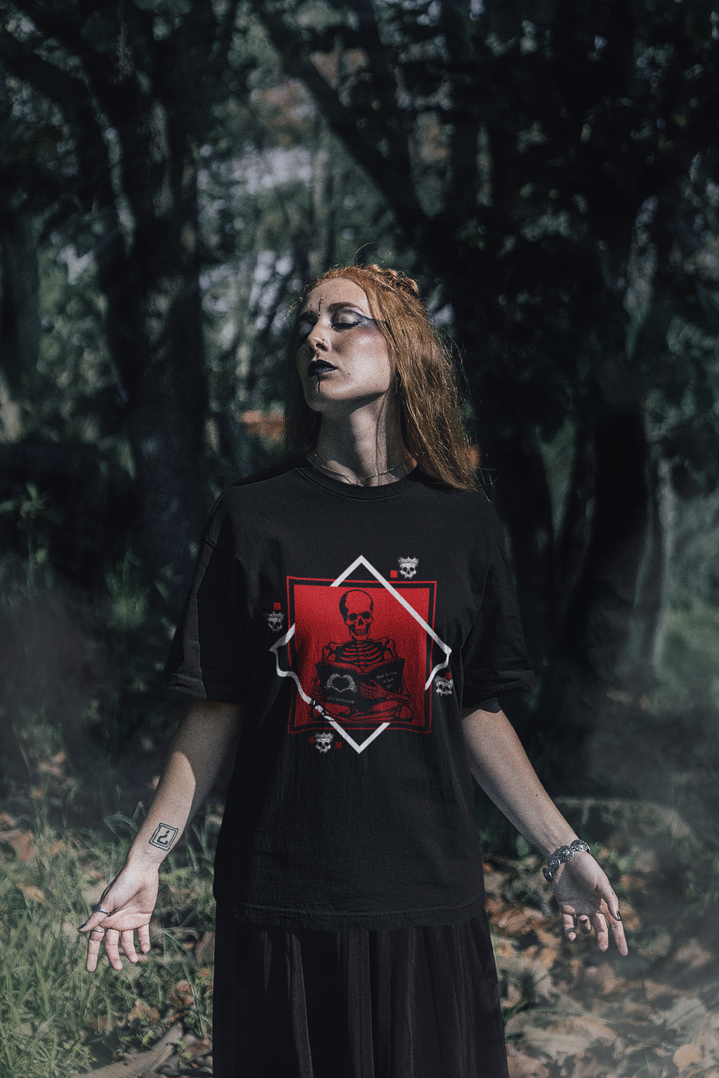 
Black t-shirt with gothic skeleton sitting cross-legged reading a book inside a framed diamond design, available in red, purple, and pink colorways