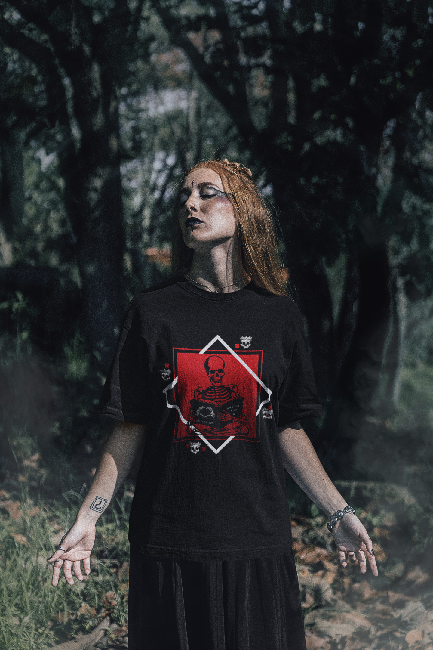 
Black t-shirt with gothic skeleton sitting cross-legged reading a book inside a framed diamond design, available in red, purple, and pink colorways